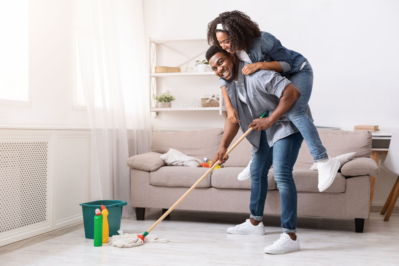 Pareja divirtiéndose mientras trapea el piso de la sala con productos de limpieza, fomentando la limpieza del hogar compartida y el bienestar.
